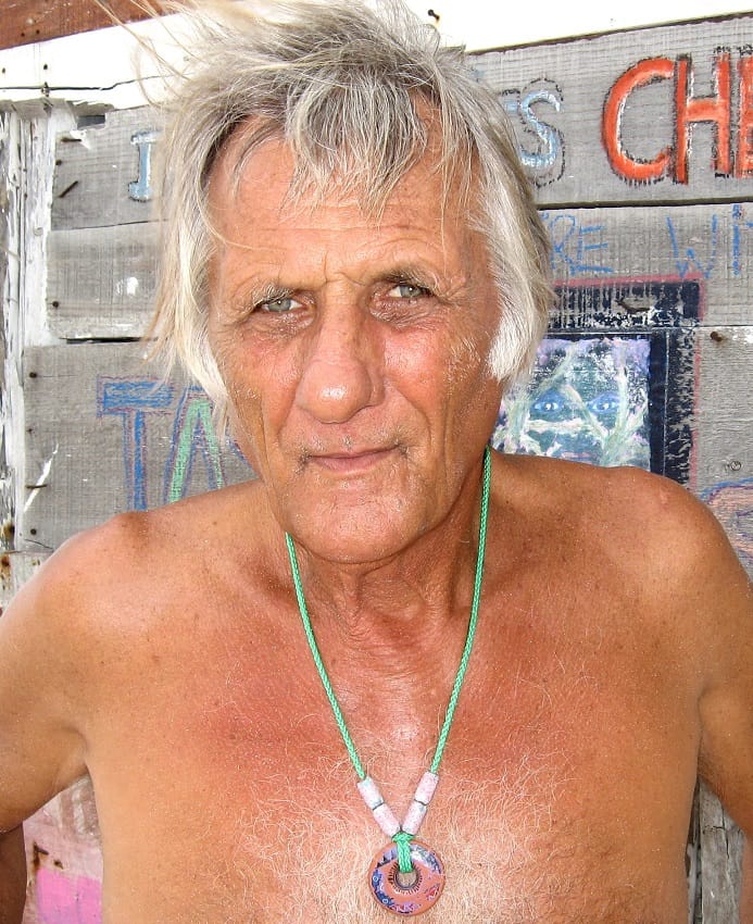 An artist looks into the camera while shirtless on a beach