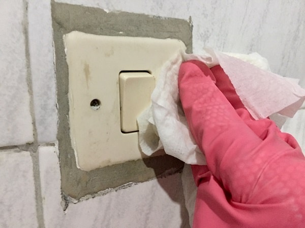 a hand in a rubber glove wipes a light switch with a disinfectant wipe from a travel cleaning kit