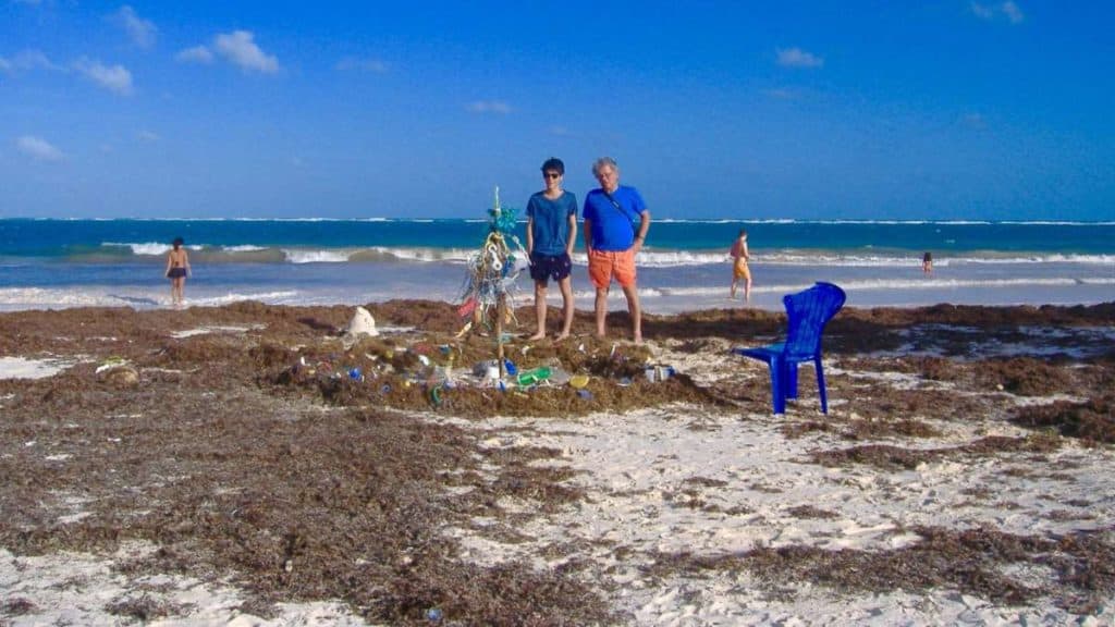man and boy stand on the beach near the water and look at an art installation made of plastic trash from the beach