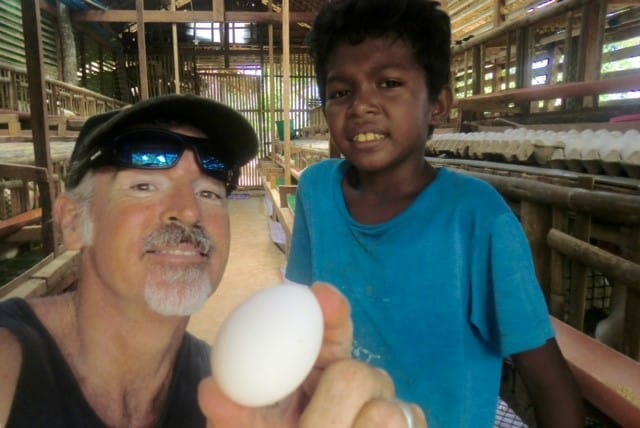Tedly holds an egg up the to the camera as he smiles next to an Ati child in the Ati chicken coop.
