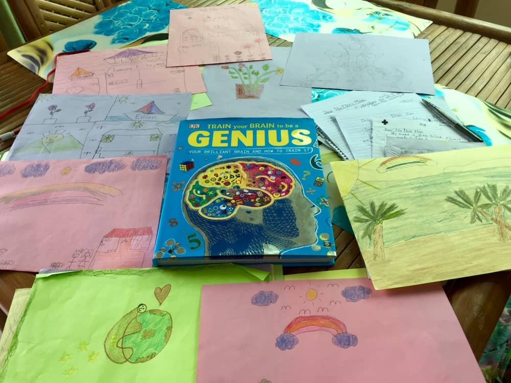 A book titled "Train your Brain to be a Genius" along with homework assignments and children's drawings spread out on a table.