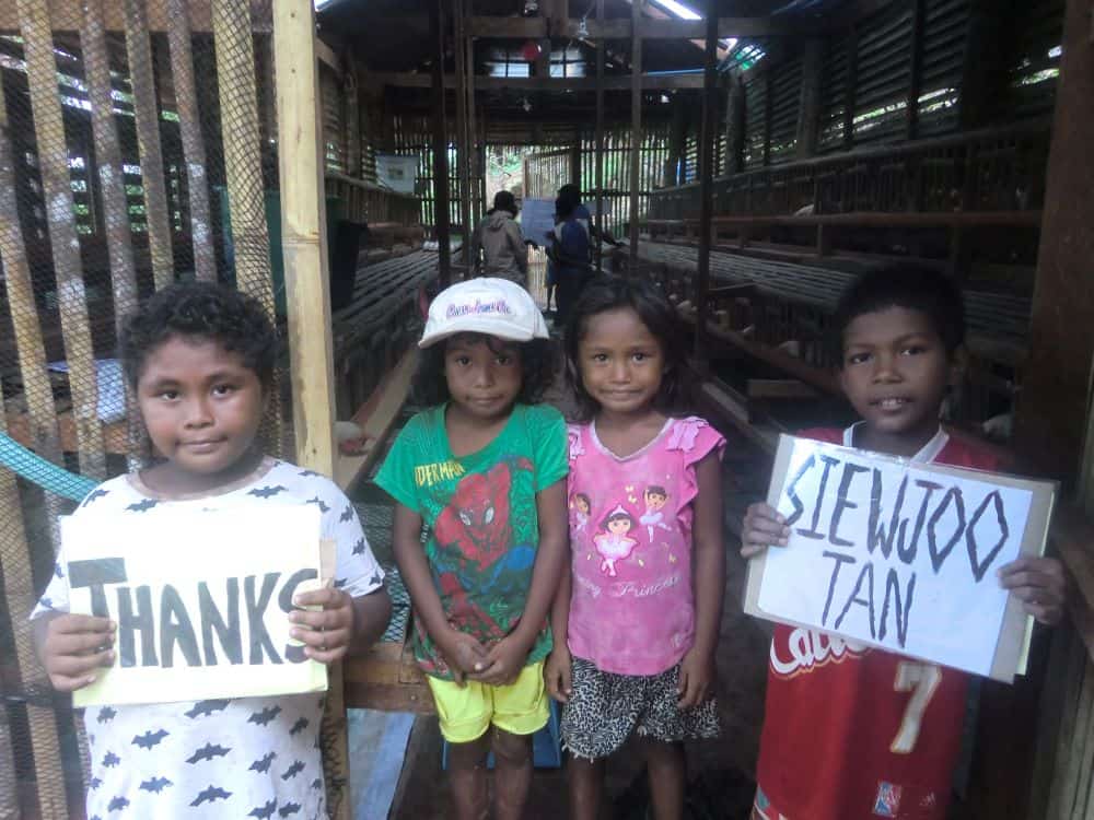 Ati children hold thank you side with chickens in the henhouse behind them.
